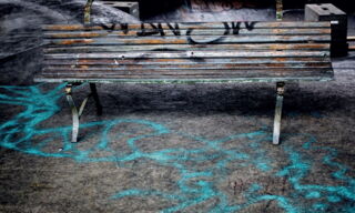 Eine kaputte Parkbank aus Holz steht mitten in einem Skaterpark, der Boden ist mit Farbe aus der Spraydose übersät.