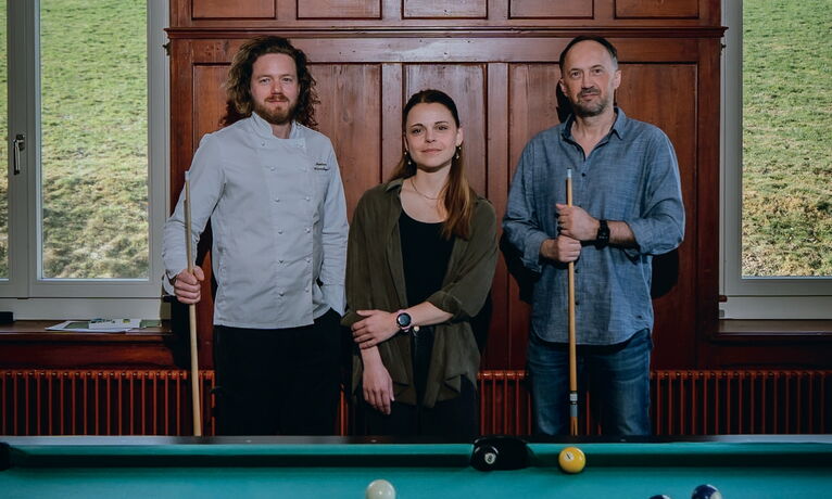 Drei Personen stehen an einem Billardtisch mit Queues. Kugeln liegen auf dem Tisch