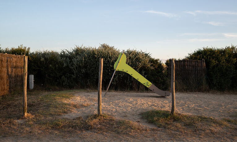 Kleine grüne Rutsche auf einem sandigen Spielplatz, umgeben von Sträuchern und Holzpfosten im warmen Abendlicht.