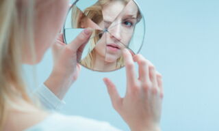 Une jeune femme blonde regarde son reflet dans un miroir brisé.