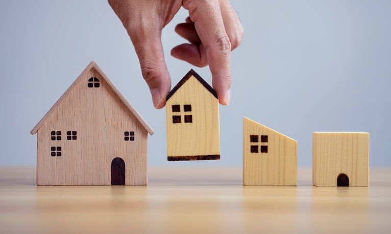 Une main tient une petite maquette en bois représentant une maison au-dessus de trois autres maisons de tailles différentes. Les maisons sont posées sur une table en bois devant un fond de couleur unie.